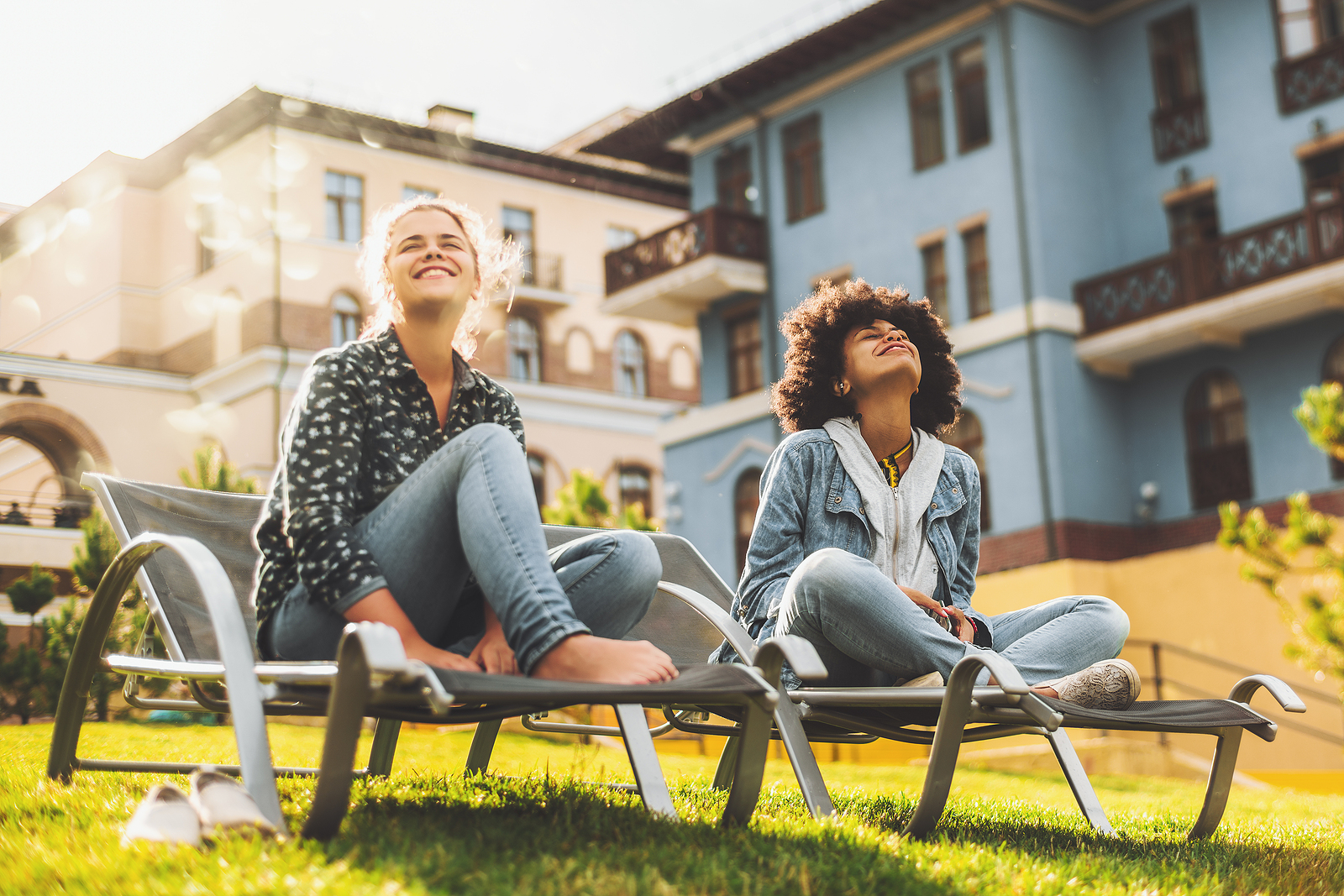 Black and Caucasian female friends on daybeds are sitting outdoors on the lawn and enjoying bright spring sun; two squinting girls of different races sitting on the recliners outdoor on the grass