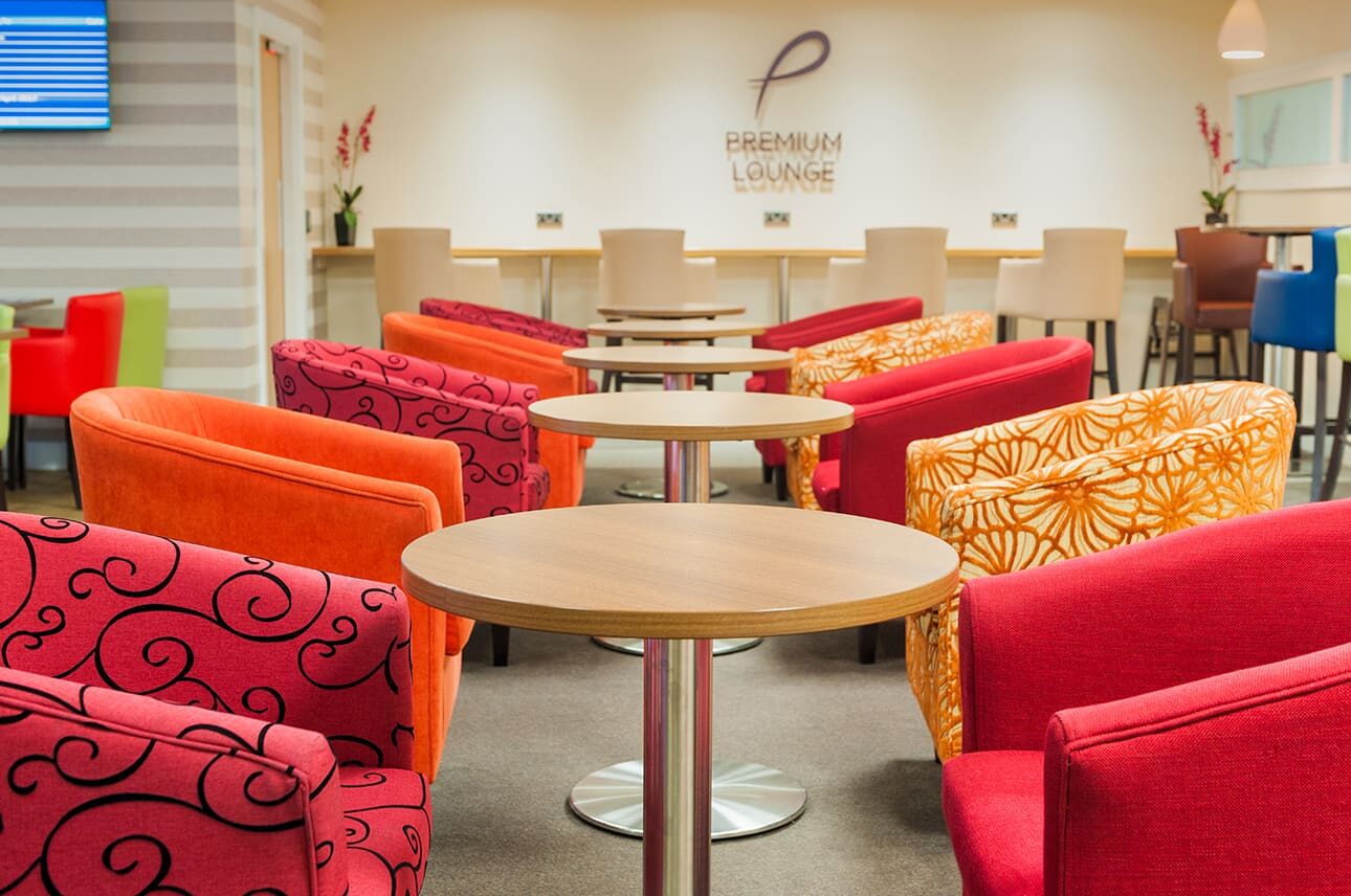 A row of tub chairs in various colours and fabrics, next to round dining tables, in a premier lounge at an airport.