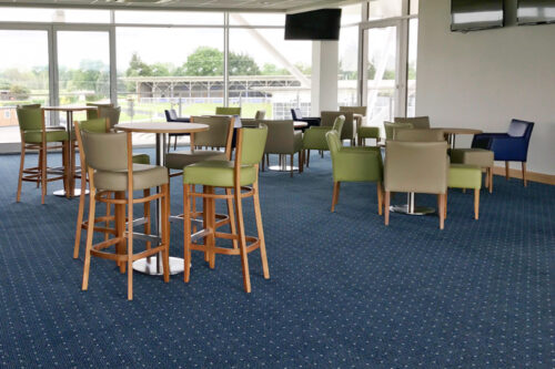 Bar stools and armchairs with round tables, at a racecourse. Blue carpet and cream walls, overlooking the race arena.