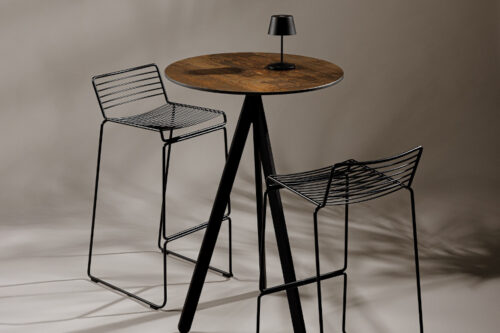 Fly folding poseur table with black legs and rust effect HPL table top paired with two black metal bar stools on a grey background.