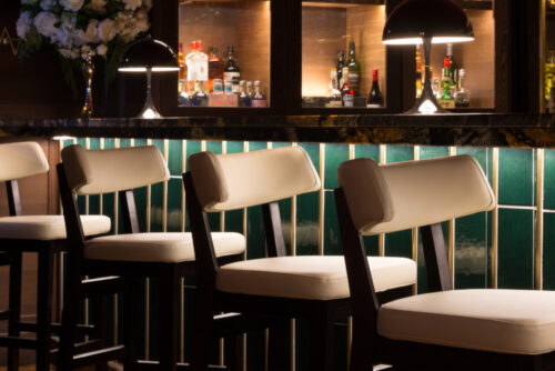 Cream bar stools next to a green tiled bar, and low level lighting.